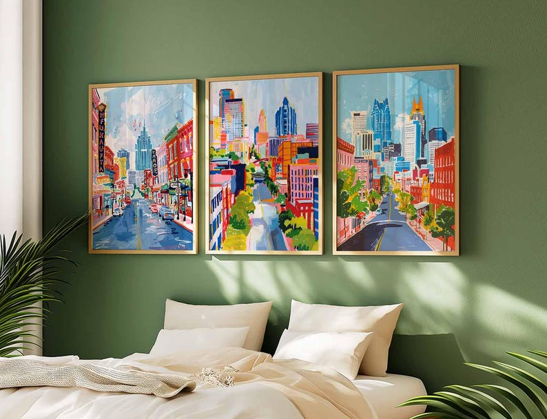 May include: Three framed cityscape paintings with gold frames hang above a bed. The artwork depicts colorful city streets with buildings, trees, and cars. The paintings are in shades of blue, red, orange, and yellow.