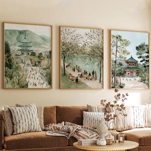 May include: Three framed watercolour prints showing Asian landscapes. The artwork features pagodas, trees, and figures, with a palette of greens, blues, and browns. The prints are displayed above a brown sofa with striped cushions.