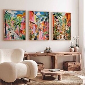 May include: Three colorful paintings of a street scene with palm trees, buildings, and people walking. The paintings are framed in wood and hung on a white wall.