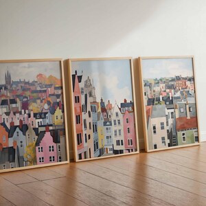 May include: Three framed art prints featuring colorful watercolor paintings of cityscapes. The artwork depicts buildings with various architectural styles and colors, including pink, yellow, and blue. The frames are made of light-colored wood.