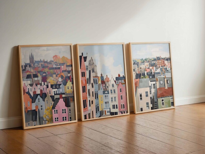 May include: Three framed art prints featuring colorful watercolor paintings of cityscapes. The artwork depicts buildings with various architectural styles and colors, including pink, yellow, and blue. The frames are made of light-colored wood.