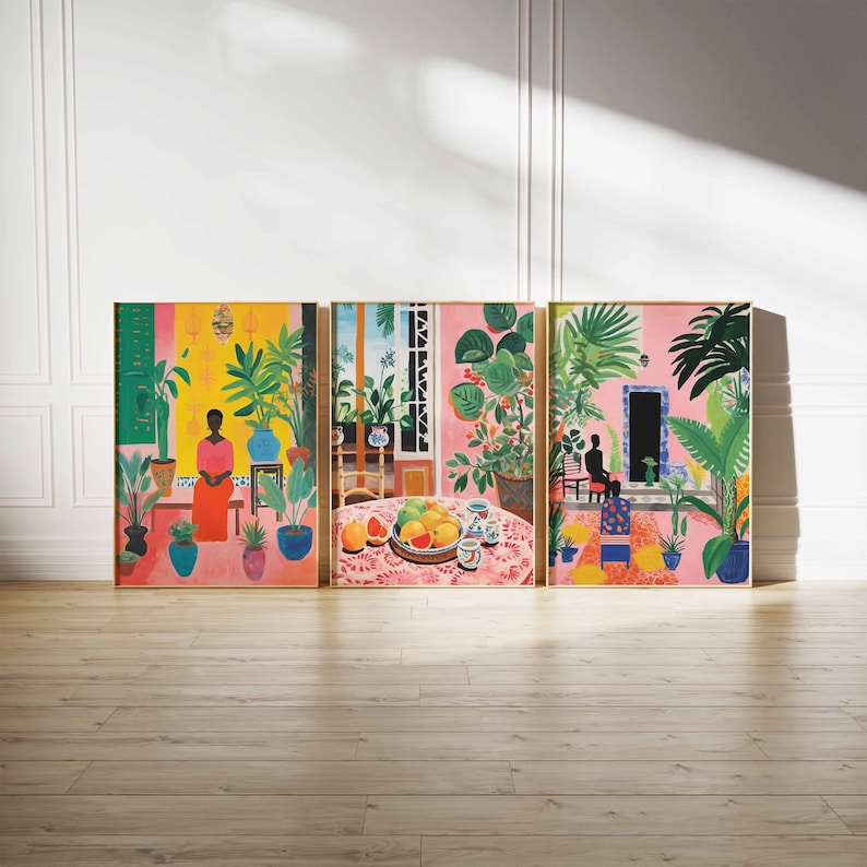 May include: Three framed art prints depicting a tropical scene with a woman in a pink dress, a table with fruit, and a silhouette of a person sitting in a chair. The prints have a gold frame and are set against a white wall.