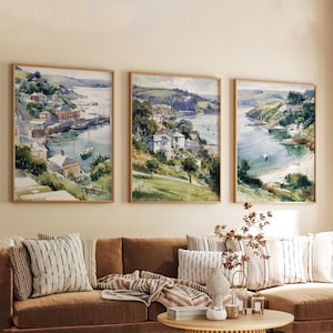 Pode incluir: Três pinturas em aquarela emolduradas retratam cenas costeiras com edifícios, barcos e vegetação exuberante. A obra de arte apresenta um porto, uma baía e um rio, todos com uma paleta de cores suaves e naturais. As pinturas são exibidas acima de um sofá marrom com almofadas listradas.