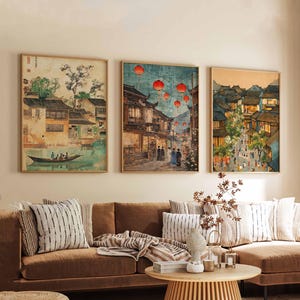 May include: Three framed prints of Japanese-style artwork depicting scenes of a city with buildings, people, and red lanterns. The prints are hung on a wall above a brown couch with pillows and a throw blanket.