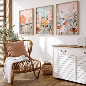 May include: Three framed illustrations of cityscapes with pastel colors and whimsical details. The illustrations are hung on a white wall above a white cabinet with a wooden top. A wicker chair with a brown cushion and a white throw blanket sits in front of the cabinet.