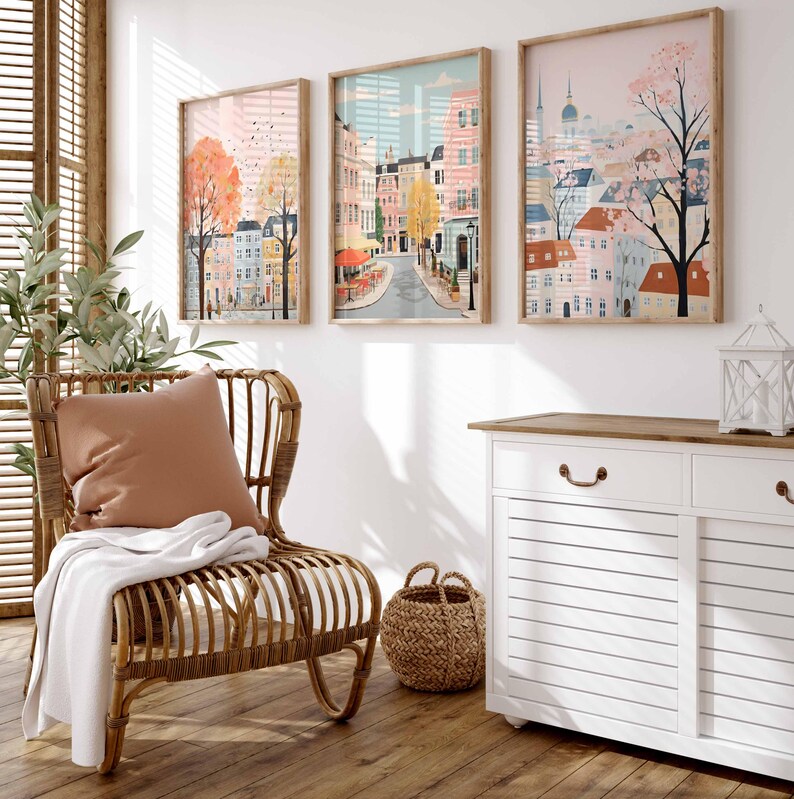 May include: Three framed illustrations of cityscapes with pastel colors and whimsical details. The illustrations are hung on a white wall above a white cabinet with a wooden top. A wicker chair with a brown cushion and a white throw blanket sits in front of the cabinet.