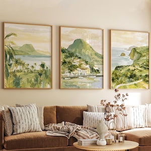 May include: Three framed watercolor landscape paintings. Each features a coastal scene with green mountains, palm trees, and buildings. The paintings are in wooden frames and hang above a brown sofa with striped pillows.