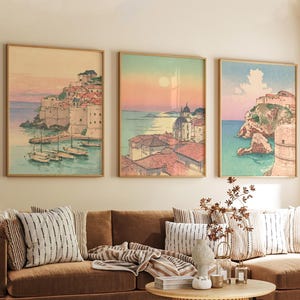 May include: Three framed art prints showcasing coastal scenes with buildings, boats, and water. The artwork has a vintage style with soft colours, including pink, blue, and beige. The prints are displayed above a brown sofa with decorative cushions.