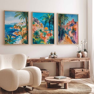 May include: Three framed prints of colorful paintings depicting a coastal town with palm trees, buildings, and a blue sky. The prints are hung on a white wall above a wooden table and a white armchair.