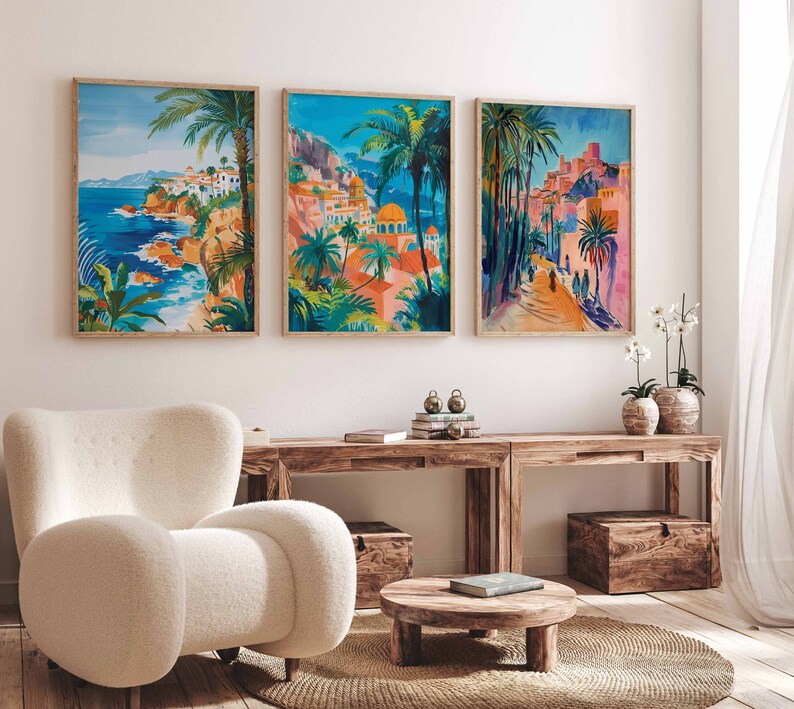 May include: Three framed prints of colorful paintings depicting a coastal town with palm trees, buildings, and a blue sky. The prints are hung on a white wall above a wooden table and a white armchair.