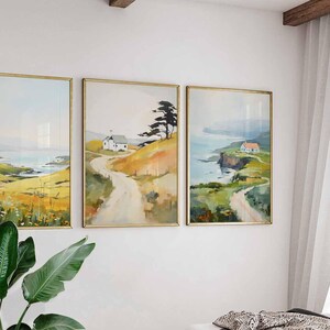 May include: Three framed landscape paintings with gold frames hang on a white wall. The paintings depict coastal scenes with rolling hills, roads, and small houses. The artwork features a soft color palette of yellow, green, and blue.