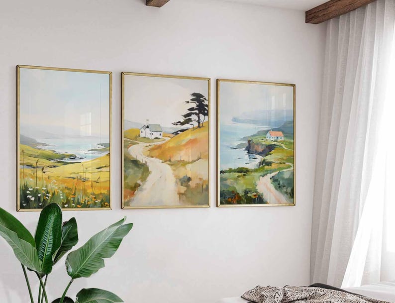 May include: Three framed landscape paintings with gold frames hang on a white wall. The paintings depict coastal scenes with rolling hills, roads, and small houses. The artwork features a soft color palette of yellow, green, and blue.