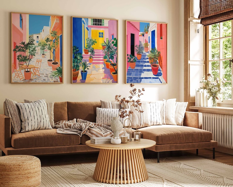 May include: A brown sectional sofa with striped throw pillows and a wooden coffee table in front of it. Three colorful paintings of a street scene with vibrant colors and potted plants hang on the wall behind the sofa.