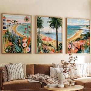Könnte beinhalten: Ein Set aus drei gerahmten Wandbildern, die Küstenszenen mit Sandstränden, Stegen, Leuchttürmen und leuchtenden Blumen im Vordergrund in Orange-, Rosa- und Grüntönen zeigen. Jeder rechteckige Druck stellt eine andere maritime Landschaft dar.