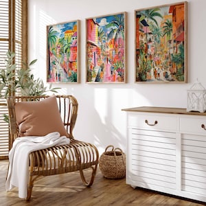May include: Three framed paintings of colorful cityscapes with palm trees and buildings. The paintings are hung on a white wall above a white cabinet with a wooden top. A wicker chair with a brown pillow and a white throw blanket sits in front of the cabinet.