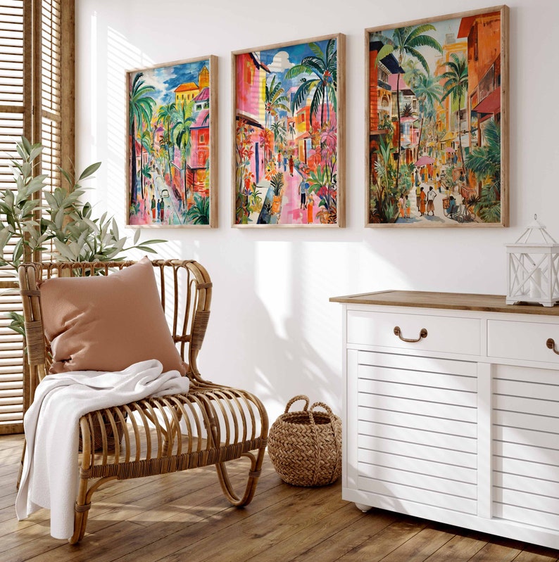 May include: Three framed paintings of colorful cityscapes with palm trees and buildings. The paintings are hung on a white wall above a white cabinet with a wooden top. A wicker chair with a brown pillow and a white throw blanket sits in front of the cabinet.