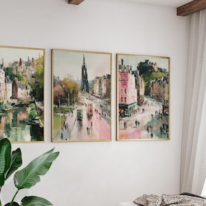May include: Three framed landscape art prints depicting city scenes with buildings, streets, and water. The artwork features a muted color palette with pink, green, and brown hues. The frames are gold-colored, and the prints are displayed on a white wall.