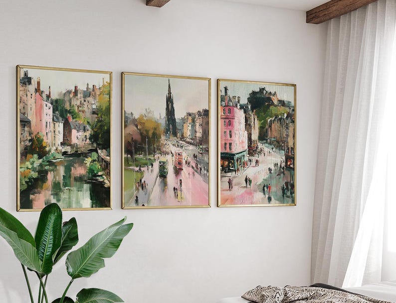 May include: Three framed landscape art prints depicting city scenes with buildings, streets, and water. The artwork features a muted color palette with pink, green, and brown hues. The frames are gold-colored, and the prints are displayed on a white wall.