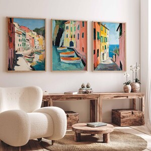 May include: Three framed paintings of colorful buildings in a coastal town. The paintings are hung on a white wall above a wooden table and a white armchair.