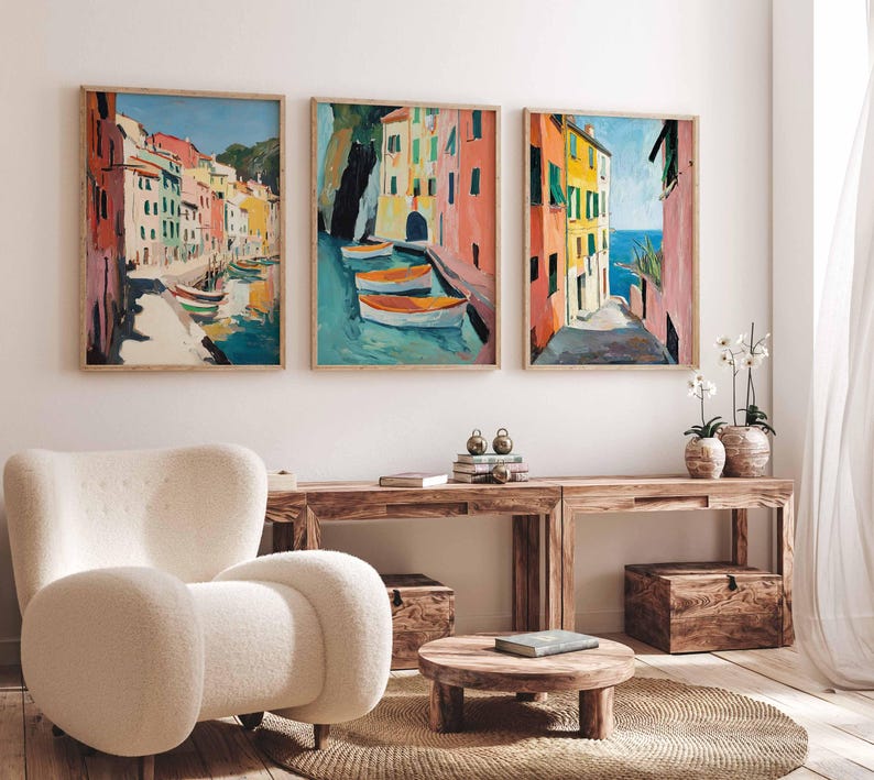May include: Three framed paintings of colorful buildings in a coastal town. The paintings are hung on a white wall above a wooden table and a white armchair.