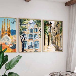 May include: Three framed art prints depicting European cityscapes. The artwork includes a colorful depiction of a cathedral, a building with blue and white details, and a narrow street with streetlights. The frames have a gold finish.