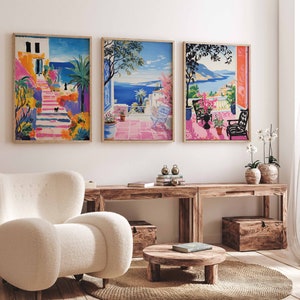 May include: Three framed art prints depicting colorful Mediterranean scenes. The artwork features vibrant depictions of coastal landscapes with buildings, stairs, and ocean views. The prints are displayed above a wooden console table with a white armchair and a round coffee table.