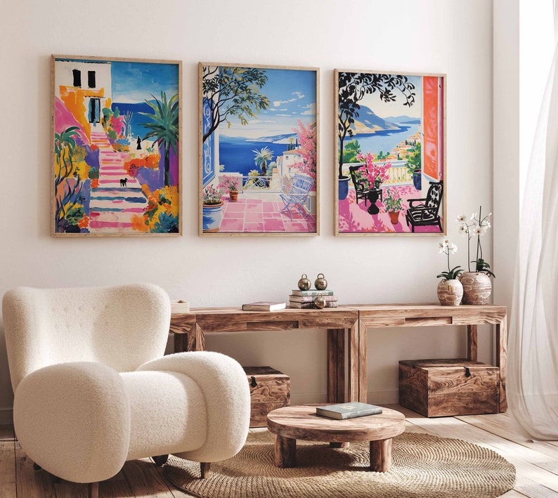 May include: Three framed art prints depicting colorful Mediterranean scenes. The artwork features vibrant depictions of coastal landscapes with buildings, stairs, and ocean views. The prints are displayed above a wooden console table with a white armchair and a round coffee table.