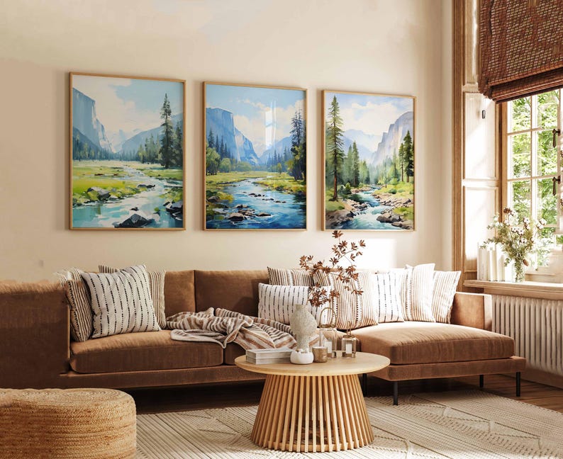 May include: Three framed landscape paintings depicting a river scene with mountains and trees. The paintings are displayed above a brown sofa with decorative pillows. A round wooden coffee table and a woven ottoman are in front of the sofa.