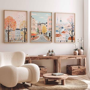 May include: Three framed illustrations of cityscapes with pastel colors and whimsical details. The first image features a pink sky with a street lined with colorful buildings and trees. The second image shows a street with a cafe and buildings in the background. The third image depicts a cityscape with a church steeple and a tree in the foreground.