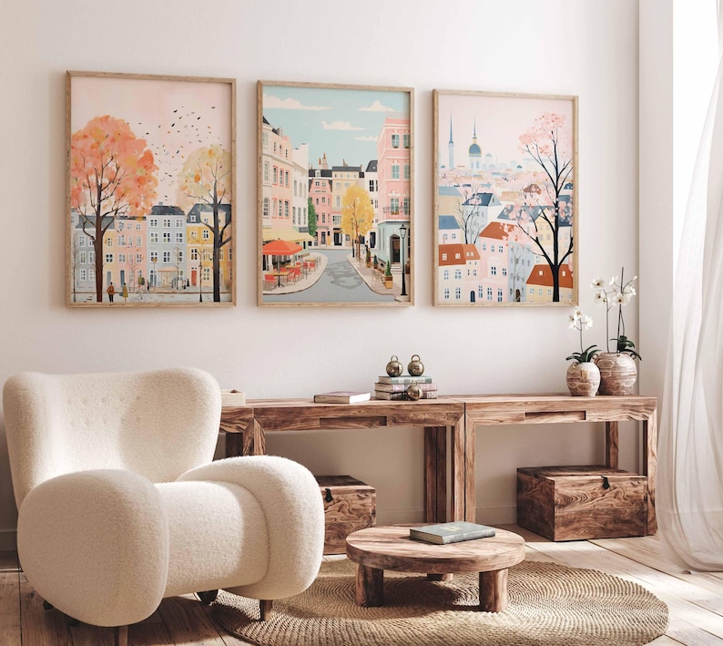 May include: Three framed illustrations of cityscapes with pastel colors and whimsical details. The first image features a pink sky with a street lined with colorful buildings and trees. The second image shows a street with a cafe and buildings in the background. The third image depicts a cityscape with a church steeple and a tree in the foreground.
