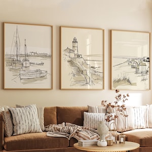 May include: Three framed art prints with coastal sketches. The first depicts boats in a harbor, the second a lighthouse on a cliff, and the third beach huts. Each print has a light beige background and a thin wooden frame.