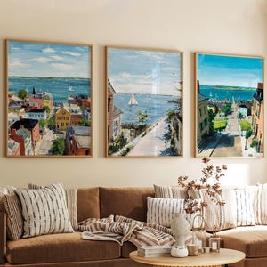Könnte beinhalten: Drei gerahmte Landschaftsbilder mit Szenen von Küstenstädten. Die Kunstwerke zeigen farbenfrohe Gebäude, ein blaues Meer mit Segelbooten und einen hellen Himmel. Die Rahmen sind hellbraun, und die Gemälde hängen über einem braunen Sofa.