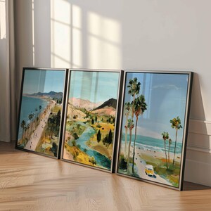 May include: Three framed landscape paintings depicting coastal and mountain scenes. The artwork features palm trees, a beach, a road, and a yellow vehicle. The paintings have black frames and are leaning against a white wall.