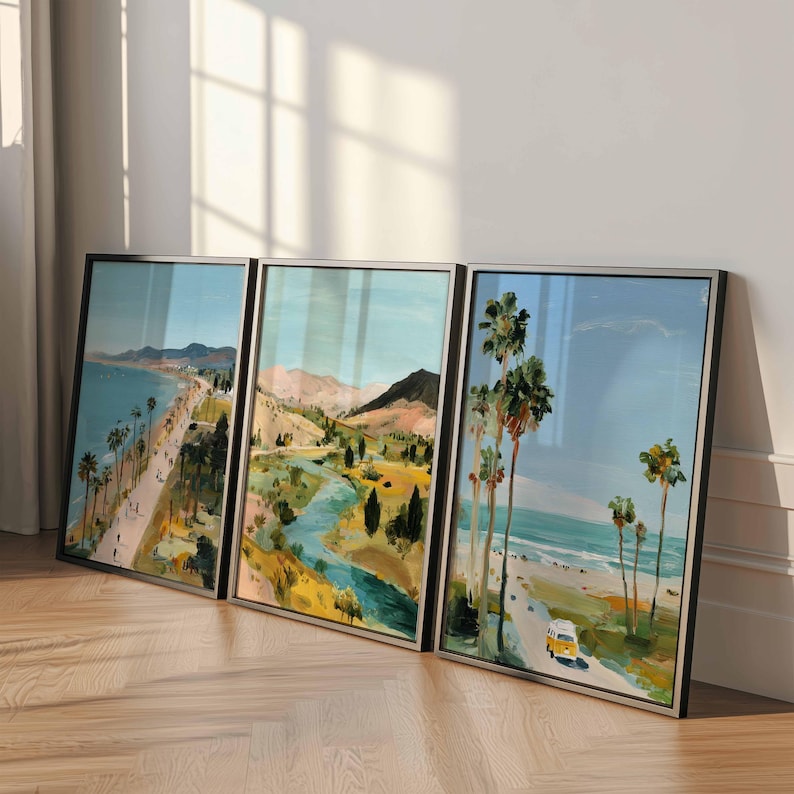 May include: Three framed landscape paintings depicting coastal and mountain scenes. The artwork features palm trees, a beach, a road, and a yellow vehicle. The paintings have black frames and are leaning against a white wall.