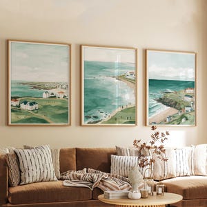 May include: Three framed watercolor prints of coastal landscapes. Each print features a different view of the ocean, beach, and cliffs, with buildings and greenery. The frames are a light wood color, and the overall color palette is muted greens and blues.
