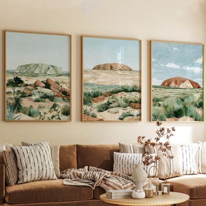 Op de afbeelding: Een set van drie ingelijste wandkunstprints met woestijnlandschappen, grote rotsformaties, schaarse groene vegetatie en een blauwe lucht. De kunstwerken bevatten aardetinten zoals bruin, beige en saliegroen in dunne houten lijsten.