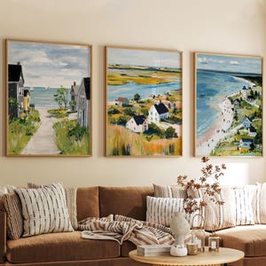 Könnte beinhalten: Drei gerahmte Landschaftsbilder mit Küstenszenen. Die Kunstwerke zeigen Häuser, Strände und Wasser, mit einer Farbpalette aus Blau, Grün und Gelb. Die Gemälde werden über einem braunen Sofa mit gestreiften Kissen ausgestellt.
