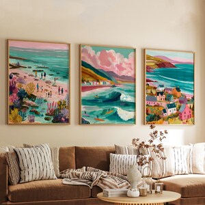 May include: Three framed art prints depicting coastal scenes. The artwork features colorful illustrations of beaches, waves, and buildings. The color palette includes shades of teal, pink, and yellow. The prints are displayed above a brown sofa with striped pillows.
