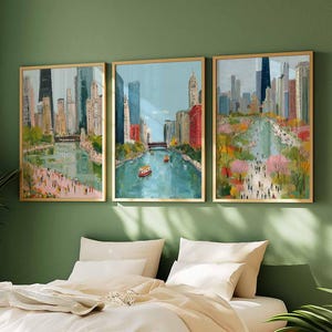 May include: Three framed art prints depicting a cityscape with a river and tall buildings. The artwork features a colorful, impressionistic style with gold frames. The paintings are displayed above a bed with white bedding, against a green wall.