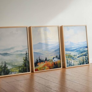May include: Three framed landscape art prints. Each print features a watercolor painting of a mountain scene with trees and a house. The frames are light-colored wood, and the artwork has a soft, muted color palette.