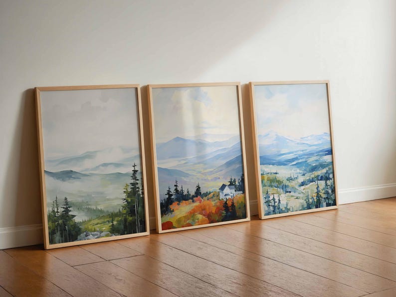 May include: Three framed landscape art prints. Each print features a watercolor painting of a mountain scene with trees and a house. The frames are light-colored wood, and the artwork has a soft, muted color palette.