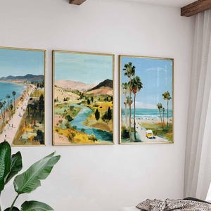 May include: Set of three framed landscape paintings. The artwork depicts coastal scenes with palm trees, beaches, and a yellow vehicle. The paintings have gold frames and are displayed on a white wall, creating a cohesive art collection.