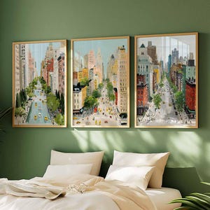 May include: Three framed watercolor paintings of a city street scene. The artwork depicts tall buildings, trees, and yellow taxis on a sunny day. The frames are a light wood color, and the paintings are hung on a green wall above a bed.