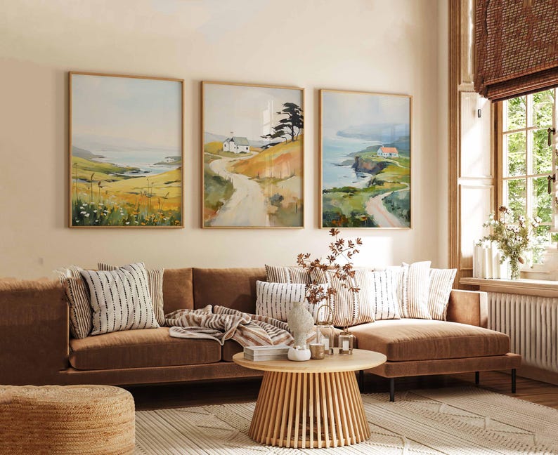 May include: Three framed landscape paintings with a coastal theme hang above a brown sofa. The paintings depict a road leading to a house, with fields and water in the background. A round wooden coffee table and patterned pillows complete the scene.