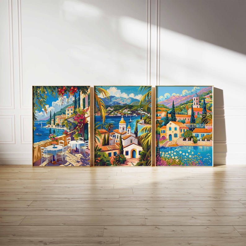 May include: Three colorful paintings depicting a Mediterranean village with white buildings, blue water, and lush green foliage. The paintings are framed in gold and hung on a white wall.