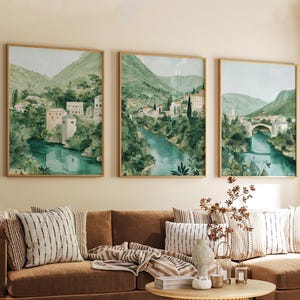 May include: Three framed watercolor landscape prints. Each print features a river flowing through a valley with buildings and lush green foliage. The frames are a light wood color, and the overall color palette is green, blue, and beige.