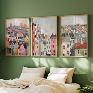 May include: Three framed watercolor prints depicting colorful townhouses and buildings. The artwork features a variety of colors, including pink, orange, blue, and gray, with gold frames. The art is displayed above a bed with white bedding and pillows.