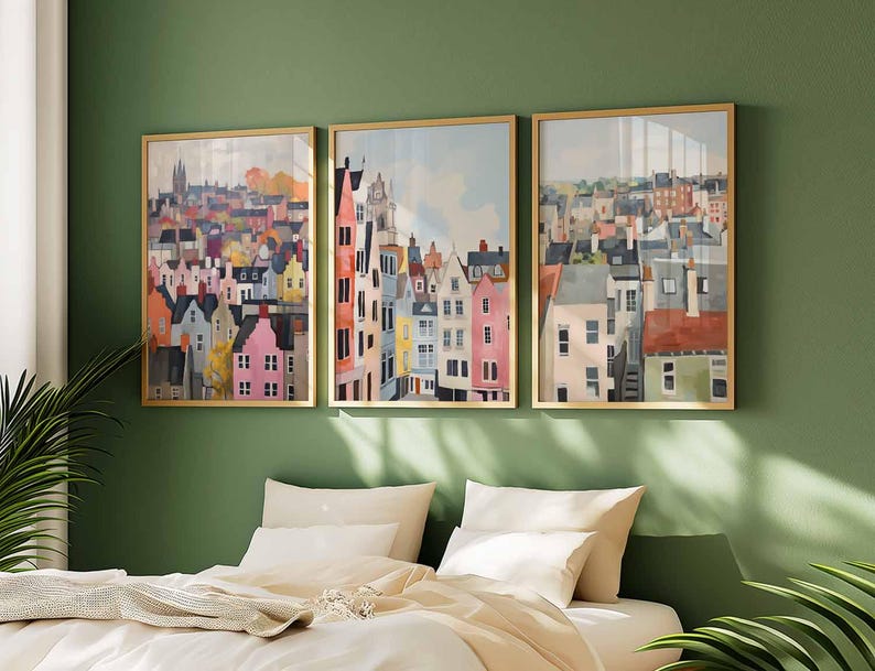 May include: Three framed watercolor prints depicting colorful townhouses and buildings. The artwork features a variety of colors, including pink, orange, blue, and gray, with gold frames. The art is displayed above a bed with white bedding and pillows.
