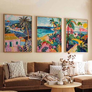 May include: Three framed paintings depicting tropical scenes with palm trees, beaches, and colorful houses. The paintings are in a living room setting with a brown couch, pillows, and a wooden coffee table.