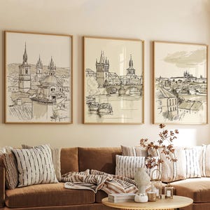 Pode incluir: Três impressões artísticas emolduradas com esboços a caneta e tinta de uma cidade europeia. A obra de arte retrata detalhes arquitetônicos de edifícios, pontes e cursos d'água. As impressões estão em molduras de madeira e penduradas acima de um sofá marrom.